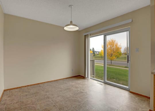 Empty room here at Westhaven Estates Apartment Homes with beige walls and a hanging light fixture. Large sliding glass doors reveal an autumn scene with a tree and grass outside.
