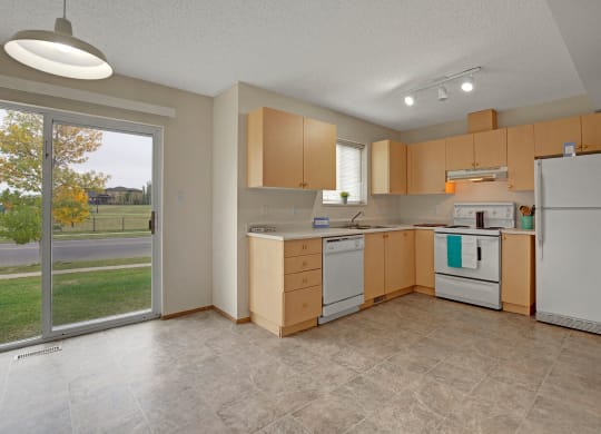 Spacious kitchen here at Westhaven Estates Apartment Homes with light wood cabinets, white appliances, and tiled flooring. A sliding glass door leads to a green lawn, evoking a light, airy feel.