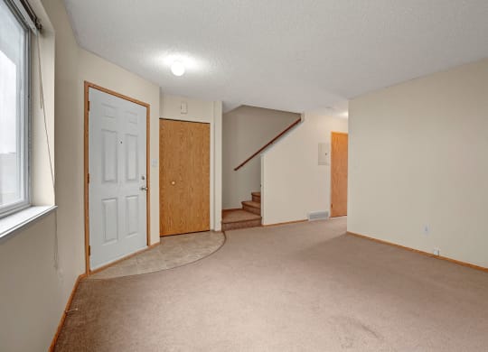 Empty living room  here at Westhaven Estates Apartment Homes with beige carpet, white walls, and a window on the left. A door and closet are near carpeted stairs leading upward, creating a calm and neutral setting.