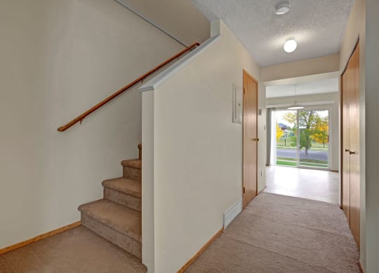 Hallway here at Westhaven Estates Apartment Homes with beige carpet and walls, featuring a staircase with a wooden railing on the left. Ahead, a bright room with a large window shows a green lawn.