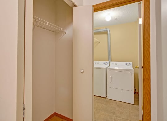 Laundry room here at Westhaven Estates Apartment Homes with beige walls and tiled floor, featuring a white washer and dryer. An open closet with a wire shelf is in the foreground.