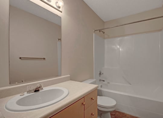 Simple bathroom here at Westhaven Estates Apartment Homes with beige walls, featuring a white sink and countertop, large mirror, toilet, and a white bathtub with a shower, under soft lighting.