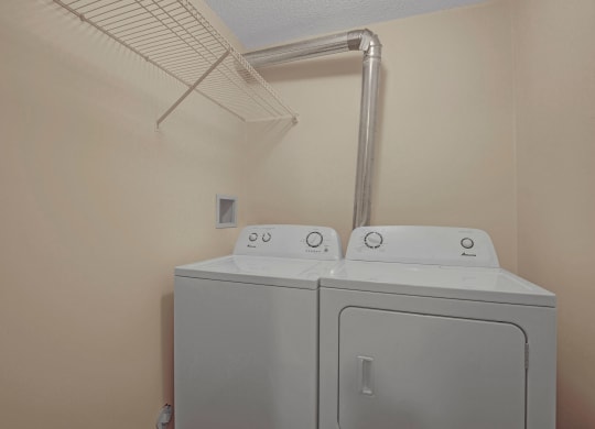 Lonely laundry room here at Westhaven Estates Apartment Homes with beige walls, white washer and dryer side by side, metal vent pipe above, and a wire shelf on the left wall.