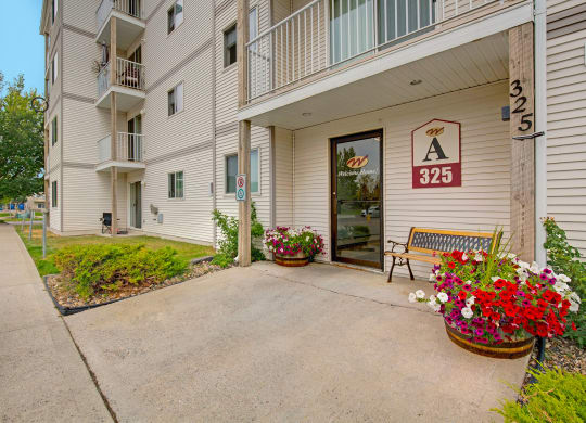 Entrance of a beige apartment building here at Westhaven Estates Apartment Homes with a "325" sign. Bright flower pots and a bench create a welcoming atmosphere. Pathway and trees are visible.
