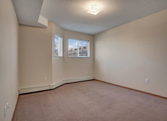 Empty room here at Windsor Terrace Apartment Homes with beige walls and carpet, and two windows showing blue sky. A ceiling light illuminates the space with a calming ambience.