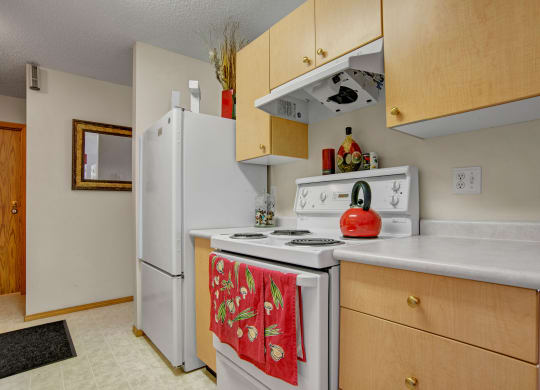 Cozy kitchen here at Windsor Terrace Apartment Homes featuring a white stove with red teapot and a red towel adorned with vegetable motifs. Light wooden cabinets add warmth.