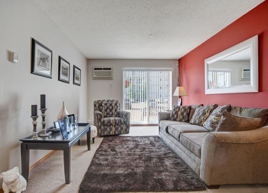 Cozy living room here at Windsor Terrace Apartment Homes with a gray couch and patterned armchair on beige carpet. A red accent wall features a large mirror. Soft light filters through vertical blinds.