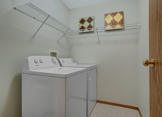 Laundry room here at Windsor Terrace Apartment Homes with a white washer and dryer side by side under a wall-mounted wire shelf. Two decorative tiles hang above the appliances. Clean and organized.