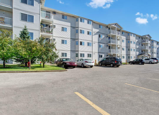 Four-story white apartment building here at Windsor Terrace Apartment Homes with balconies set in a sunny day. Cars are parked in front, and trees add greenery to the scene.