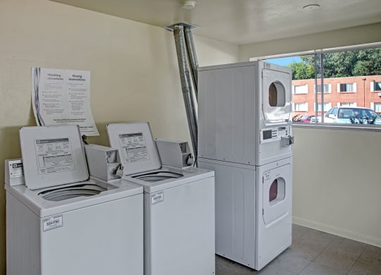 A laundromat here at Bristol Square Apartment Homes with two top-loading washers and a stacked dryer. Instruction sheets hang on the wall, and a window shows parked cars and a building.