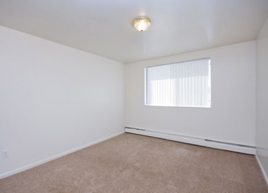 Empty room here at Bristol Square Apartment Homes with white walls, beige carpet, and a ceiling light fixture. A window with vertical blinds allows natural light, creating a neutral, calm tone.