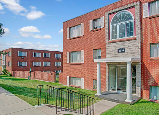 Red brick Bristol Square Apartment Homes buildings under a blue sky, with large windows and white trim. A grassy area and sidewalk lead to a columned entrance marked "1510".