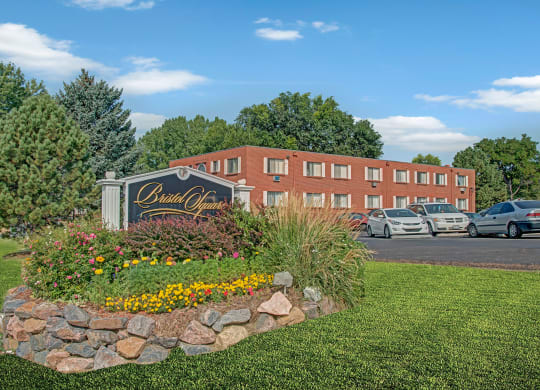 Manicured garden with colorful flowers and a "Bristol Square" sign in front. A brick residential building with parked cars is visible behind, under clear blue sky.