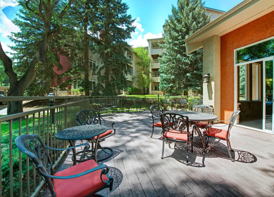 Outdoor patio here at Cheyenne Creek Apartment Homes with wrought iron tables and red-cushioned chairs in a sunny garden. Tall trees provide shade, creating a serene and inviting atmosphere.