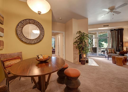 Stylish apartment interior here at Cheyenne Creek Apartment Homes with a circular dining table, wicker chairs, wall mirror, and potted plant. Bright, cozy vibe with large windows and light fixture.