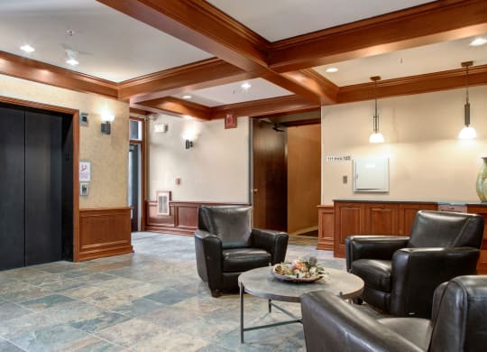 Lobby here at Cheyenne Creek Apartment Homes with modern decor, featuring leather chairs, a stone-tiled floor, and a round table with a centerpiece. An elevator and wooden trimming add warmth.