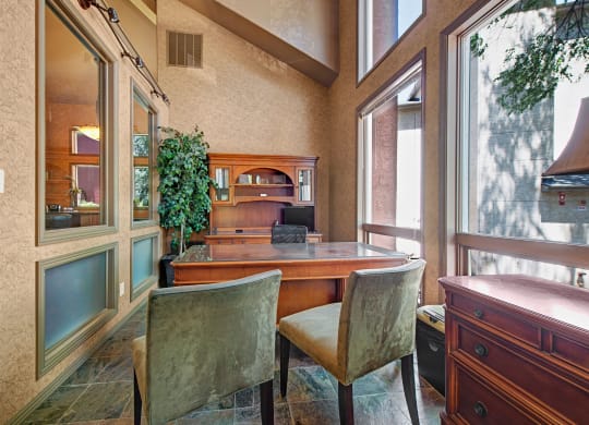 Sunlit office here at Cheyenne Creek Apartment Homes with a wooden desk, two beige chairs, a large potted plant, and tall windows. The atmosphere is calm and professional.