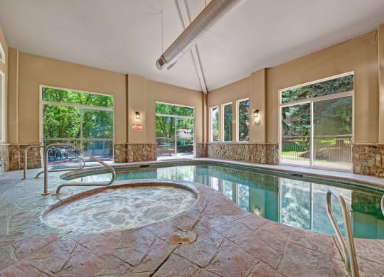 Indoor pool area here at Cheyenne Creek Apartment Homes with a bubbling jacuzzi in the foreground and a swimming pool beside it. Large windows show lush green trees outside, creating a serene atmosphere.