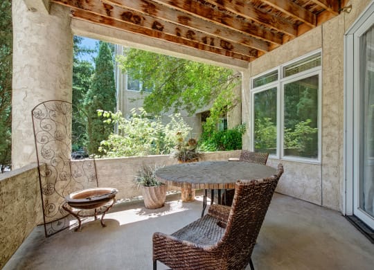 A serene patio here at Cheyenne Creek Apartment Homes with a rustic round table and two wicker chairs. Sunlight filters through overhead beams, and lush greenery surrounds the space, creating a tranquil ambiance.
