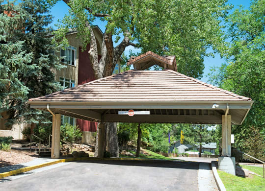 Covered driveway entrance here at Cheyenne Creek Apartment Homes with a shingled roof, surrounded by lush green trees and a larger building in the background. Calm, welcoming atmosphere.