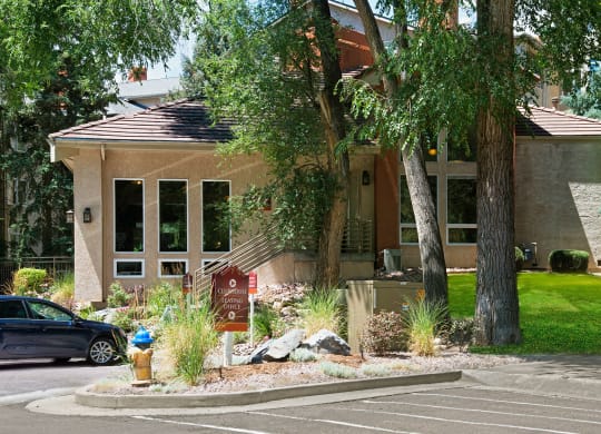 A beige building here at Cheyenne Creek Apartment Homes with large windows and a brown tiled roof is surrounded by trees and lush greenery. A sign reads "Clubhouse Leasing Office."