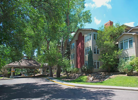 A residential building here at Cheyenne Creek Apartment Homes with large windows is surrounded by lush trees and greenery under a clear blue sky. The scene is serene and inviting.