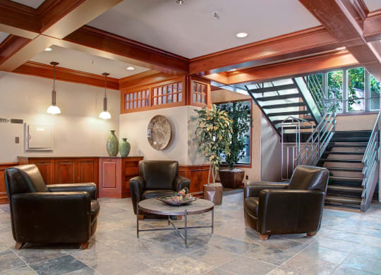 Cozy lobby here at Cheyenne Creek Apartment Homes with four black leather chairs around a round coffee table. The room has tile flooring, wood accents, a staircase, and a large potted plant.