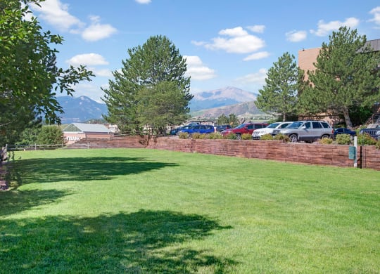 Lush green lawn here at Eagle Ridge Apartment Homes with scattered trees and mountain backdrop under a blue sky. A row of parked cars lines a wooden fence in the distance. Calm and serene.