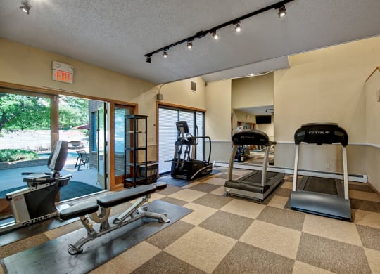 Small gym here at Eagle Ridge Apartment Homes with checkered flooring features treadmills, a recumbent bike, and a weight bench. Large windows to the left show a green outdoor area.