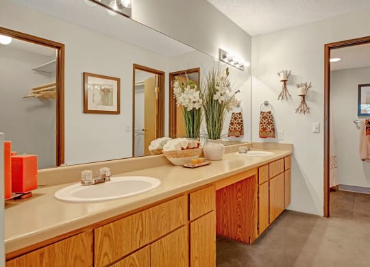 Spacious bathroom here at Eagle Ridge Apartment Homes with twin sinks, wood cabinets, and beige countertop. Decorative plants and towels add elegance, creating a serene, clean atmosphere.