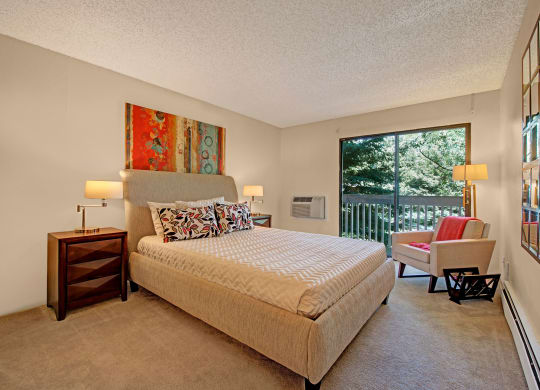 Contemporary bedroom here at Eagle Ridge Apartment Homes with a beige bed, colorful abstract art, and large window overlooking trees. Warm lighting and a cozy armchair add comfort.