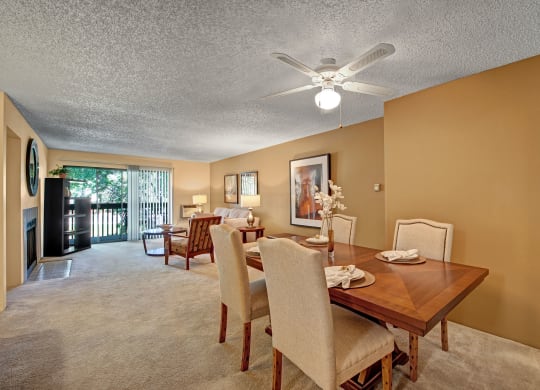 A cozy living-dining area here at Eagle Ridge Apartment Homes with beige walls and carpet. A wooden dining table is set for four. A sofa and coffee table face a large sliding glass door, allowing natural light. A ceiling fan gently illuminates the space, evoking warmth and comfort.