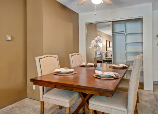 Cozy dining room here at Eagle Ridge Apartment Homes with a wooden table set for four. Beige upholstered chairs surround the table, which is decorated with plates, napkins, and a vase of white flowers. Neutral tones create a warm, inviting atmosphere.