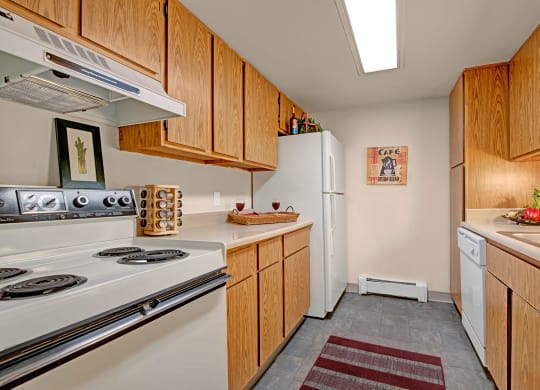 Narrow kitchen here at Eagle Ridge Apartment Homes with wooden cabinets, white appliances, and a light grey floor. A stove is on the left, a refrigerator in the center, and a dishwasher on the right. A tray with wine glasses and a fruit bowl add warmth.
