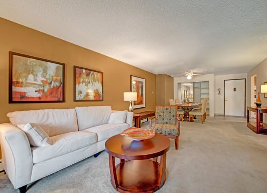 A cozy living room here at Eagle Ridge Apartment Homes with a white sofa, abstract paintings on a tan wall, and a round wooden coffee table. The dining area is in the background, with soft lighting creating a warm, inviting atmosphere.