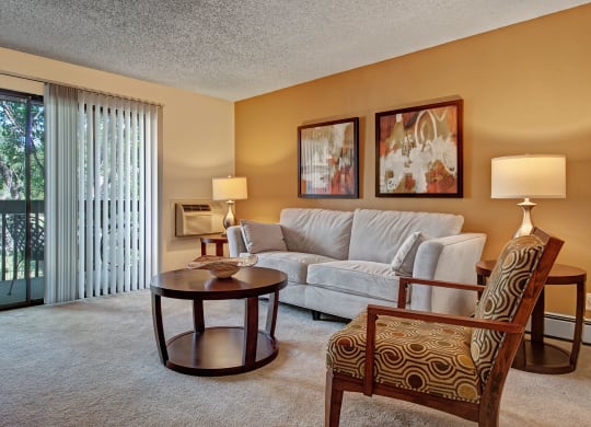 Modern living room here at Eagle Ridge Apartment Homes with beige walls, a gray sofa, and abstract art above. Two lamps illuminate the space. Glass doors reveal trees outside. Cozy and inviting.