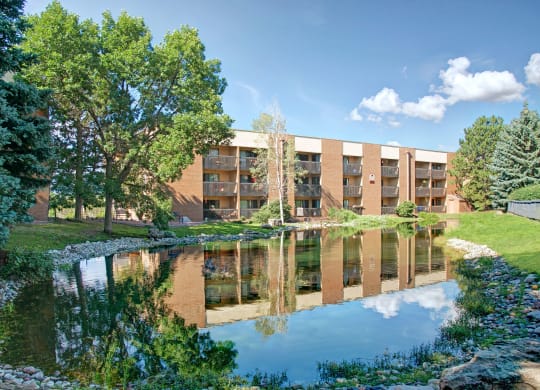 A serene pond here at Eagle Ridge Apartment Homes reflects a four-story brick apartment building surrounded by lush green trees under a blue sky with fluffy clouds, conveying tranquility.
