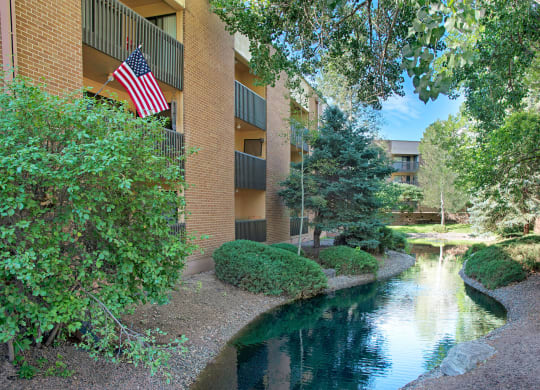 Eagle Ridge Apartment Homes building with balconies, displaying an American flag. A calm stream runs through lush greenery and trees, creating a serene atmosphere.