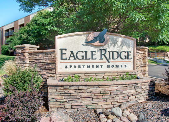 Stone sign for "Eagle Ridge Apartment Homes" with eagle silhouette, surrounded by greenery and trees, set in a landscaped area near a road.