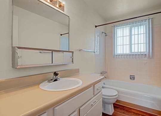 Bright bathroom here at Yorkshire Square Apartment Homes with a beige counter, sink with metallic faucet, mirror above, bathtub with tiled walls, and a window with vertical blinds.
