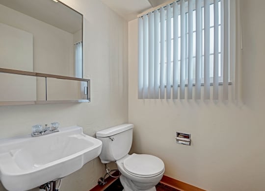 Small bathroom here at Yorkshire Square Apartment Homes with a white sink, chrome faucet, toilet, and wall-mounted mirrored cabinet. Vertical blinds cover the window, creating a clean, minimalist vibe.