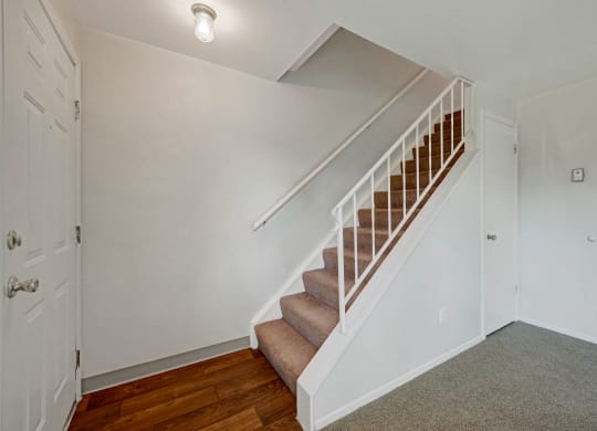 Simple hallway here at Yorkshire Square Apartment Homes with a brown carpeted staircase, white railing, and adjacent white walls. Warm lighting and wooden floor create a welcoming atmosphere.