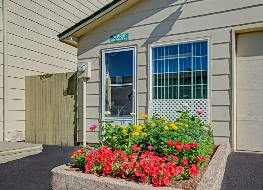 A small beige leasing office here at Yorkshire Square Apartment Homes with flowers in front, including red and yellow blooms. Bright, sunny day with a clear blue sky. The mood is cheerful.