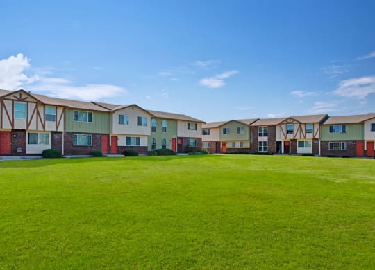 Row of suburban Yorkshire Square Apartment Homes with red doors and Tudor-style accents, surrounded by a large, well-kept grassy lawn under a clear blue sky.