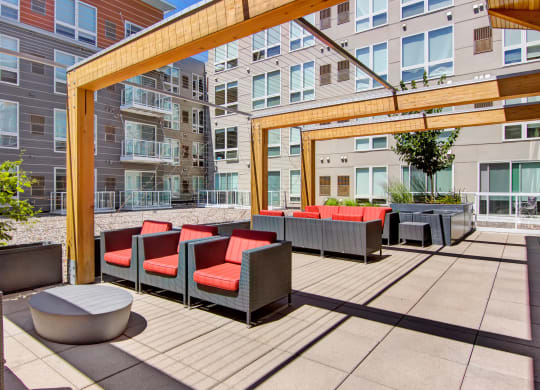 Modern apartment courtyard here at 7west Apartment Homes with gray buildings, featuring wooden pergolas and red-cushioned patio furniture. Bright, sunny atmosphere with a relaxed vibe.