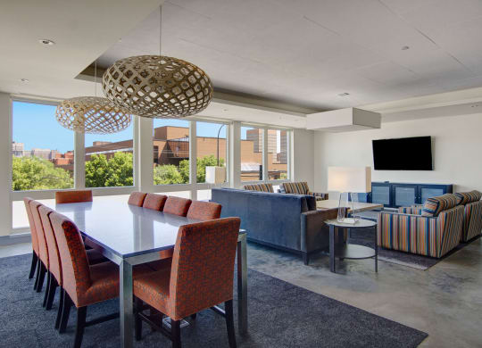 Modern open-concept resident lounge here at 7west Apartment Homes with large windows. Includes a dining table with red chairs, blue sofas, striped armchairs, and a wall-mounted TV.