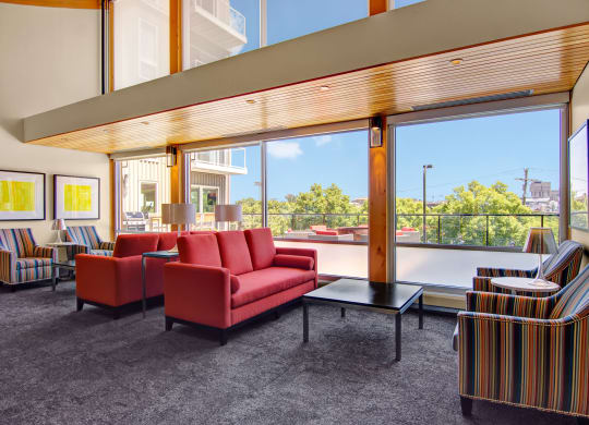Spacious modern lounge here at 7west Apartment Homes with large windows, bright red couches, striped chairs, and abstract yellow art. Natural light creates a vibrant, welcoming atmosphere.