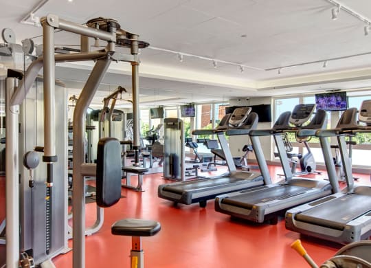 Modern gym here at 7west Apartment Homes with red flooring, featuring treadmills, weight machines, and large windows allowing natural light. Energetic and inviting atmosphere.
