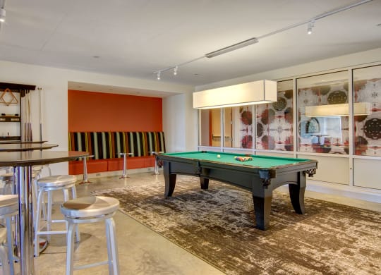 Modern recreation room here at 7west Apartment Homes with a pool table on a patterned rug. There's a seating area with striped cushions, high tables with stools, and abstract wall art.