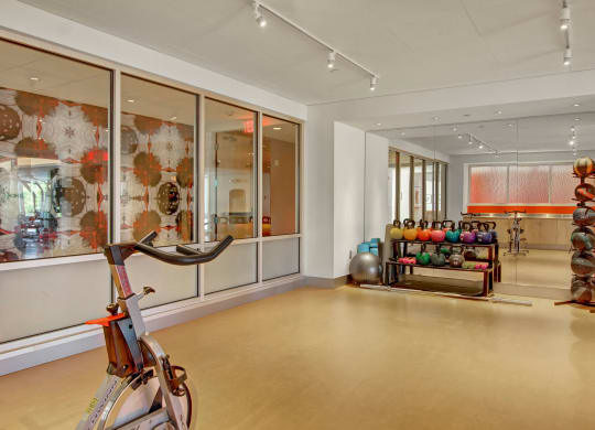 Bright gym room here at 7west Apartment Homes with exercise bikes, colorful kettlebells, medicine balls, and a stability ball. Modern decor includes abstract art on glass walls.